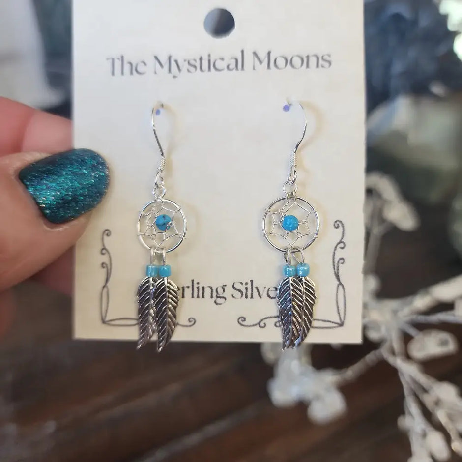 Sterling silver earrings featuring an intricate dreamcatcher with two feather charms, displayed on a white background. Perfect for protection, inspiration, and boho spirit.