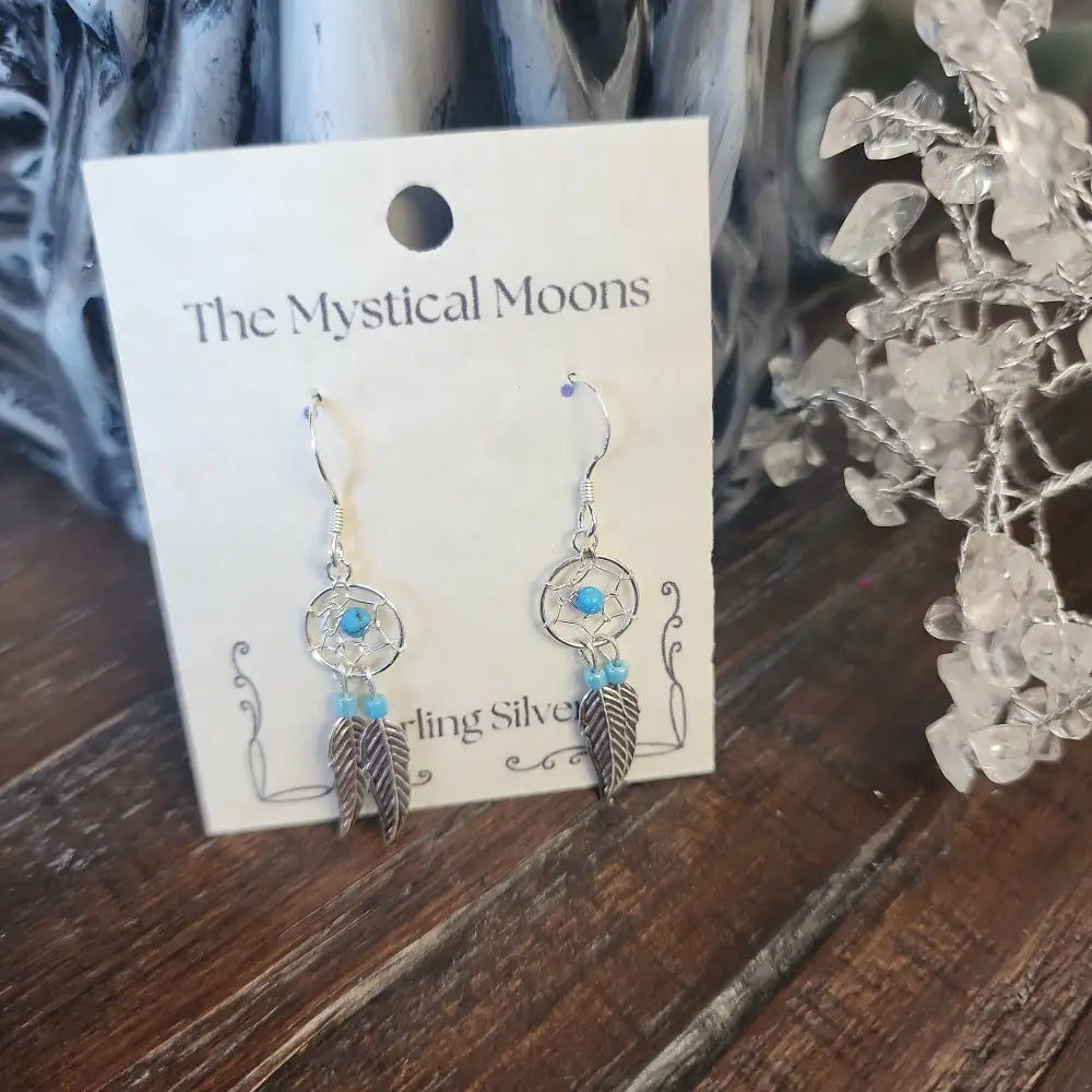 Sterling silver earrings featuring an intricate dreamcatcher with two feather charms, displayed on a white background. Perfect for protection, inspiration, and boho spirit.