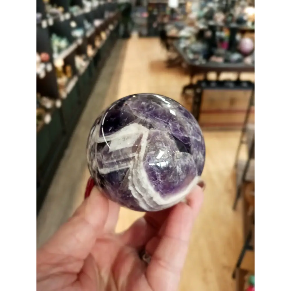 Hand-polished Dream Amethyst sphere featuring vivid purple and white chevron bands, displayed on a white background.