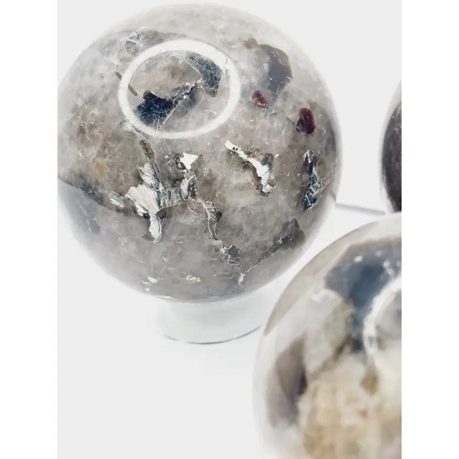Quartz, garnet, tourmaline, and mica sphere, polished with clear, red, black, and shimmering inclusions, displayed on a white background.
