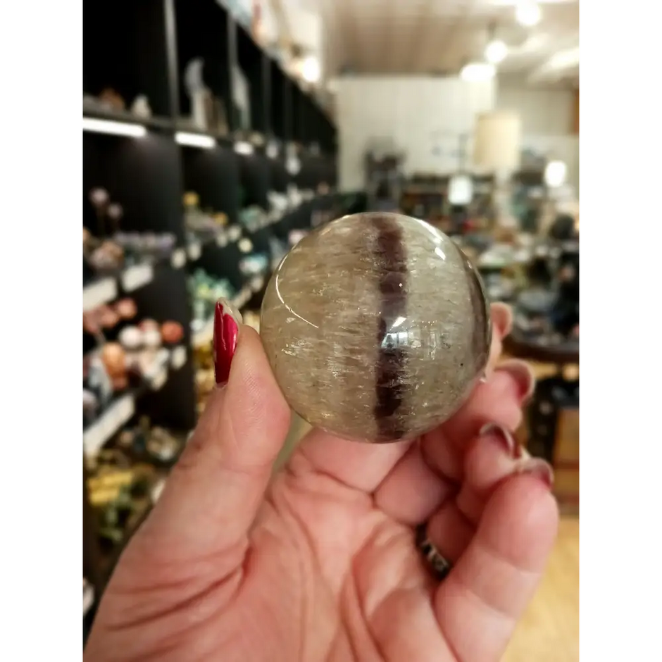 Hand-polished Rainbow and Clear Fluorite sphere featuring luminous bands of purple, green, blue, and clear tones, displayed on a white background.
