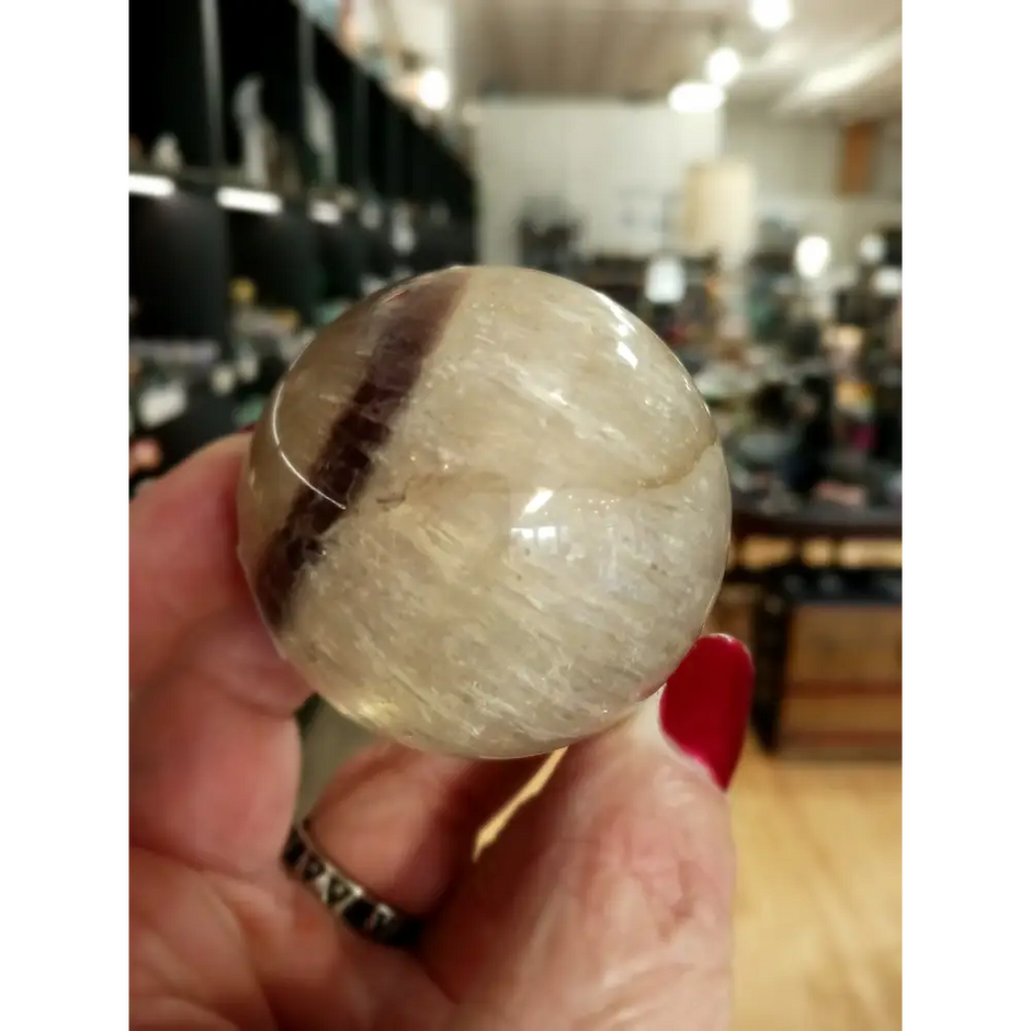 Hand-polished Rainbow and Clear Fluorite sphere featuring luminous bands of purple, green, blue, and clear tones, displayed on a white background.