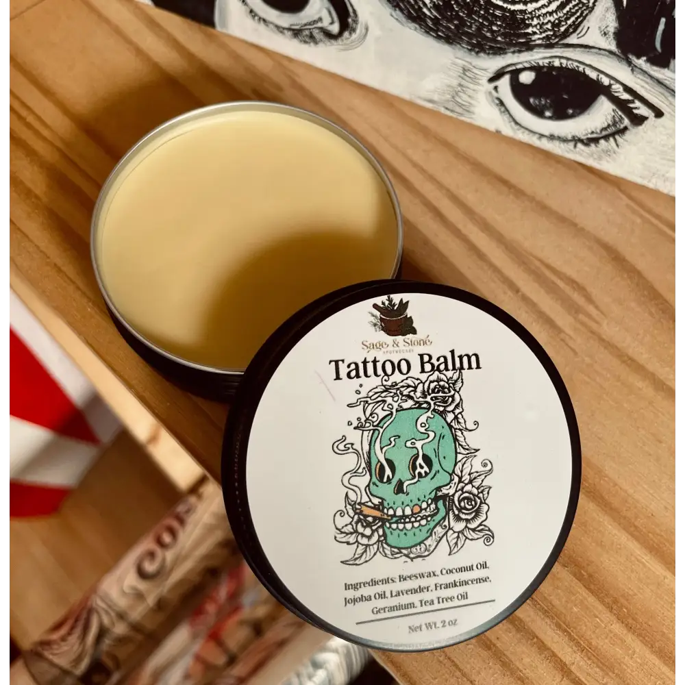 Tattoo Healing Balm in a small jar, surrounded by fresh herbs and natural oils, displayed on a white background. Perfect for soothing, protecting, and revitalizing tattoos.

